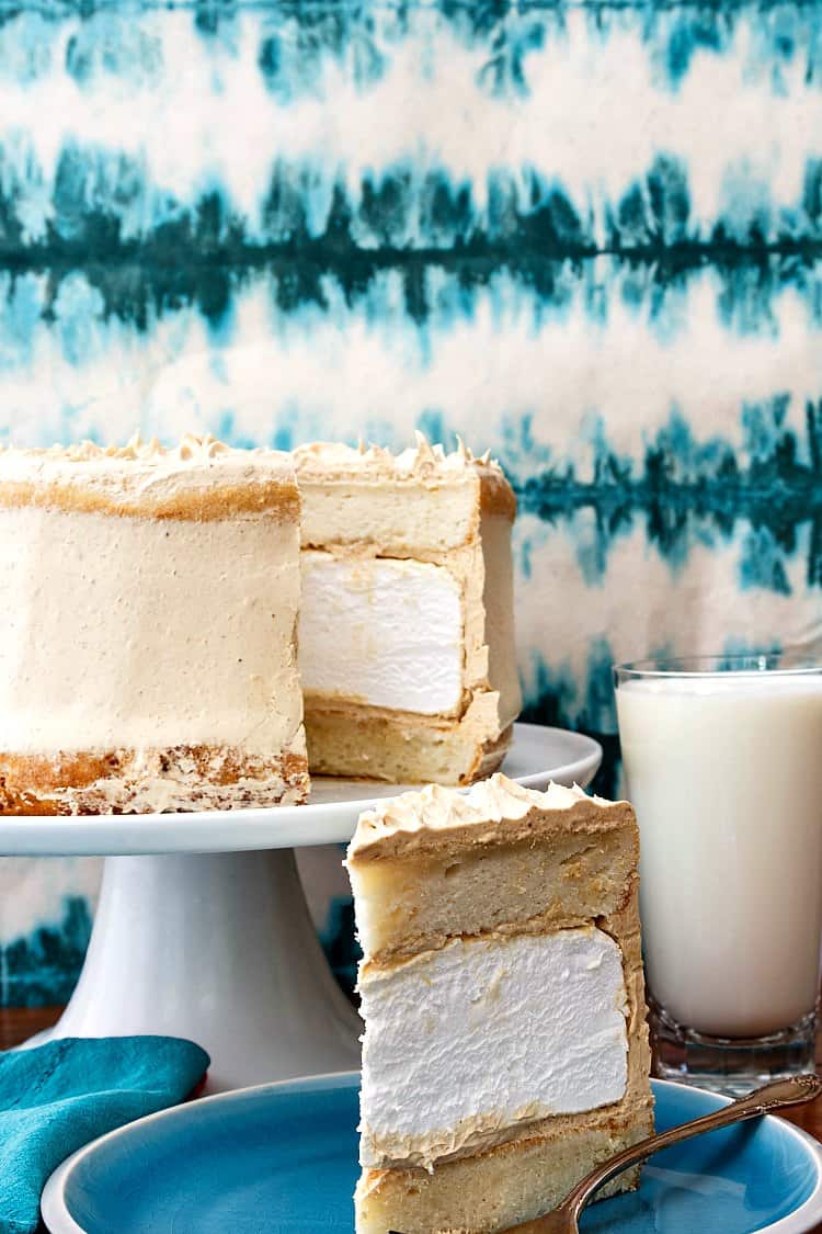 Layer cake with white cake layers, homemade marshmallow, and peanut butter buttercream on a blue plate. One slice is removed showing interior of cake. The slice is on a plate next to a glass of milk.