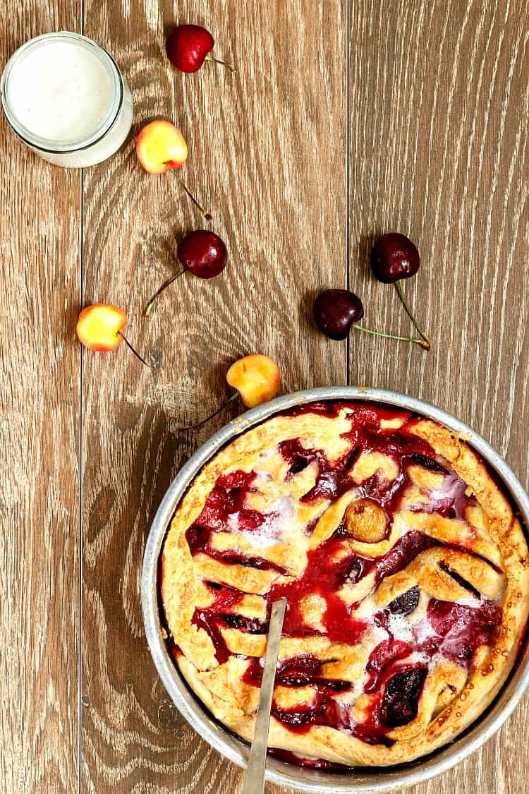 A round tin of deep dish of baked cobbler or sonker with some whole cherries scattered around on a wooden background.