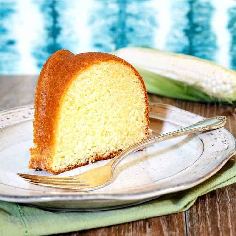 A slice of buttermilk pound cake on a plate with a fork and a fresh ear of corn in the background.