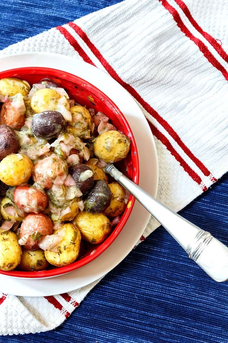 My no mayo potato salad made with whole baby red, white, and blue potatoes in a red bowl on a white plate with a blue placemat as the background.