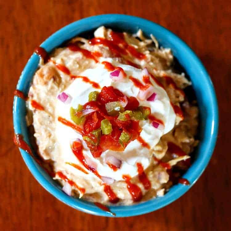 An overhead shot of Keto Crack Chicken in a blue bowl and topped with sour cream, diced tomatoes and jalapenos and a zig zag of hot sauce.