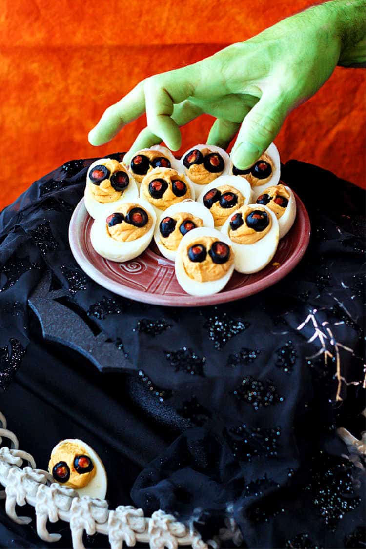 A platter of devilish Halloween deviled eggs on a black spider web pedestal with a green hand reaching for one.