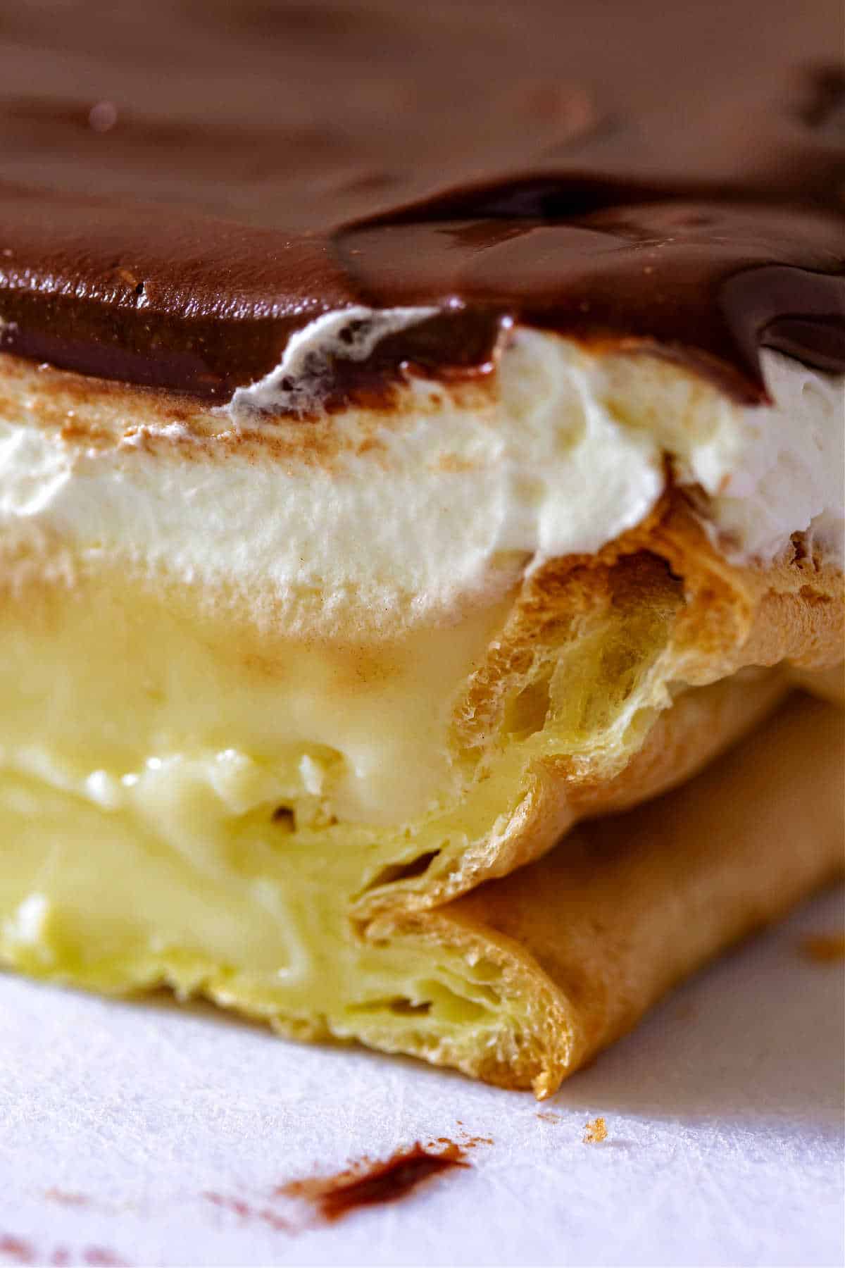 A close-up of a slice of cream puff cake showing the layers of choux paste, vanilla custard, whipped cream and chocolate ganache.