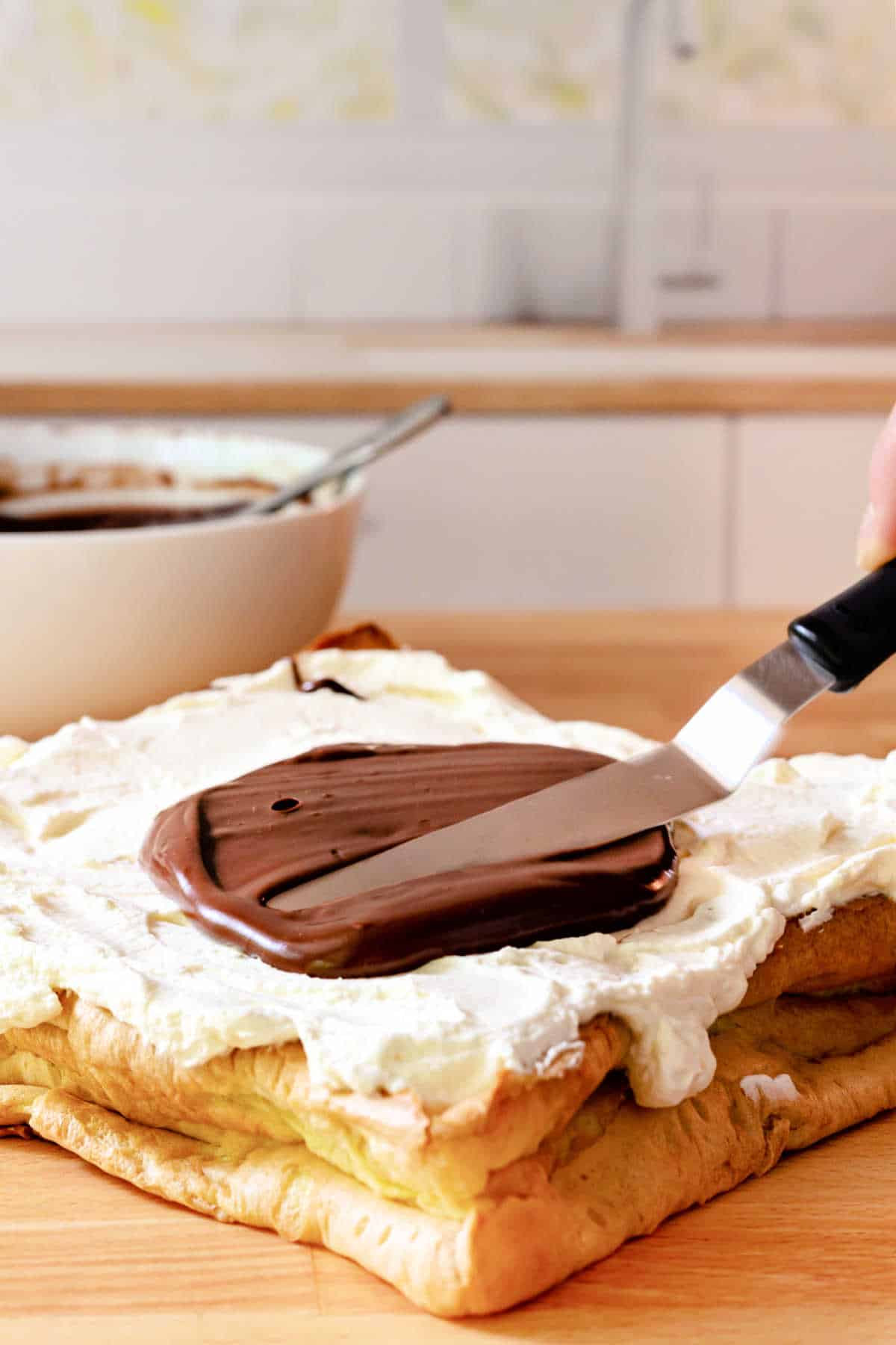A square pastry spread with whipped cream. A small spatula is spreading chocolate ganache over the whpped cream, and there is a white bowl of ganache in the background.