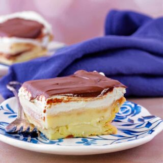 A slice of layered eclair dessert on a blue flowered plate with a blue napkin and another slice of cream puff cake in the background.