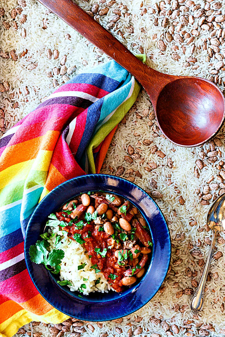 Overhead shot of a wide-rimmed blue bowl with rice and beans, topped with salsa on a background of dried rice and beans with a ladle, napkin and spoon.