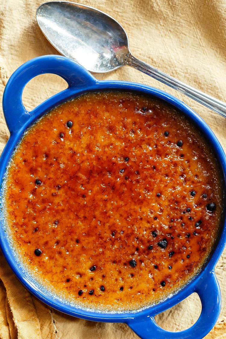 Overhead shot of blue ramekin with handles with deeply caramelized creme brulee and a silver spoon.