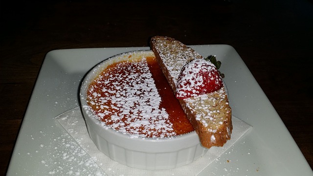 White ramekin of creme brulee with biscotti, a half strawberry and powdered sugar ready for serving.