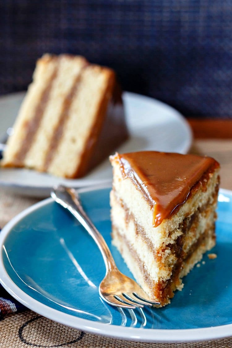 3 layer slice of butterscotch cake upright on a blue plate with a slice of the same cake on its side in the background.