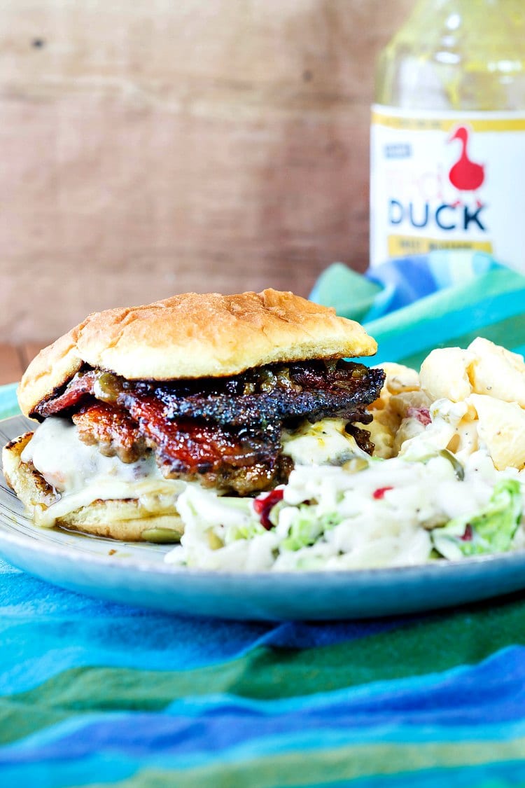 Double bacon bbq burger on a potato bun with a bottle of bbq sauce in the background.