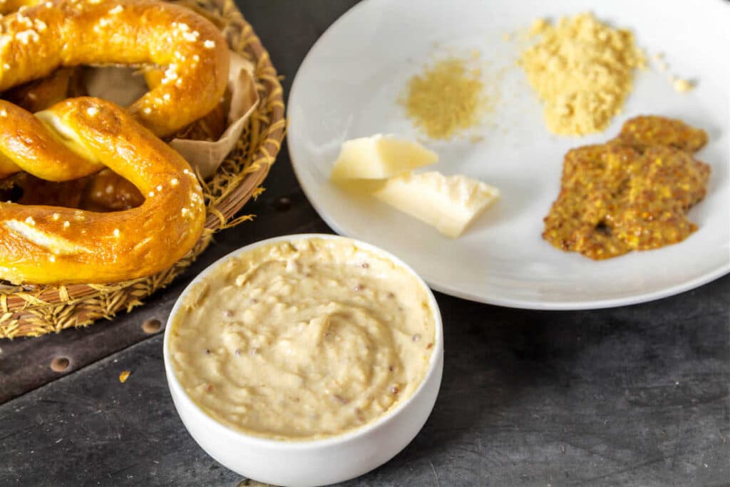 A small white bowl of beige dip, a white plate with dried spices on it, and a soft pretzel in a basket.