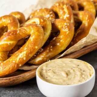 A small white bowl with cheese dip in it in front of a tray of soft pretzels.