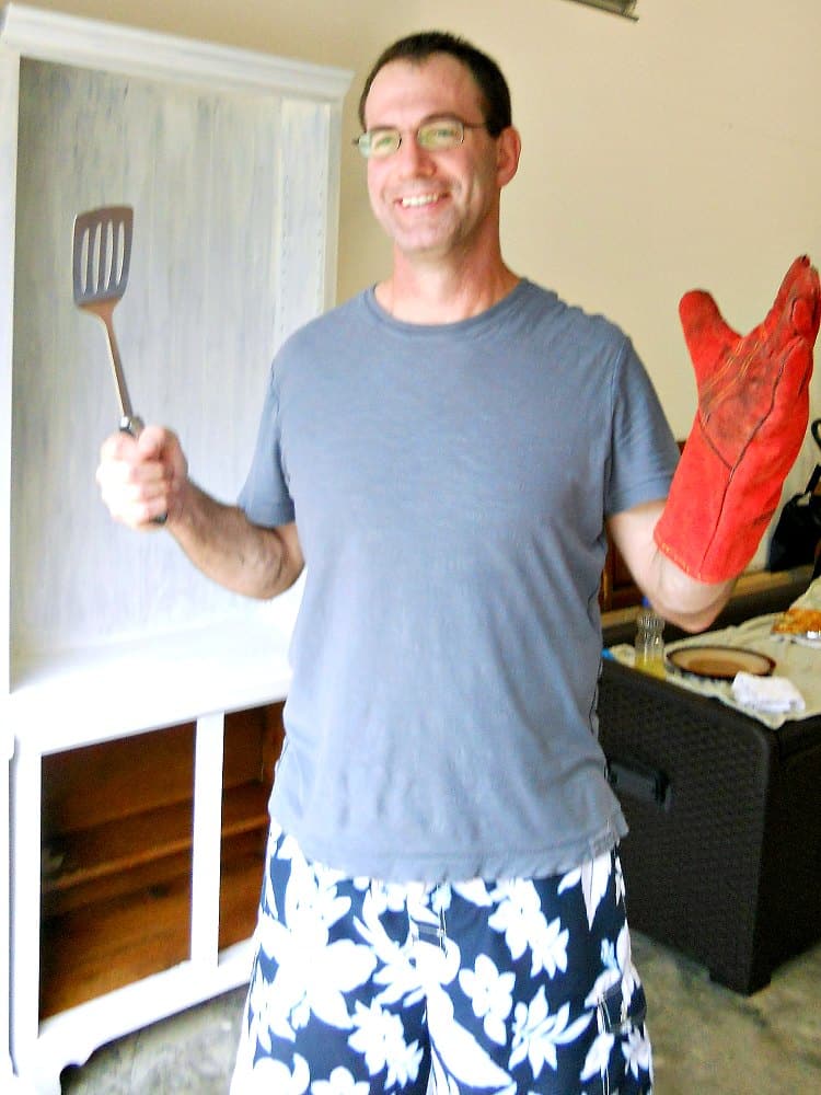A man wearing white and blue print shorts and a blue tee shirt. He is wearing wire-rimmed glasses and smiling. In one hand he is holding the handle of a spatula, and his other arm is covered to the elbow in a long, red suede welding glove.