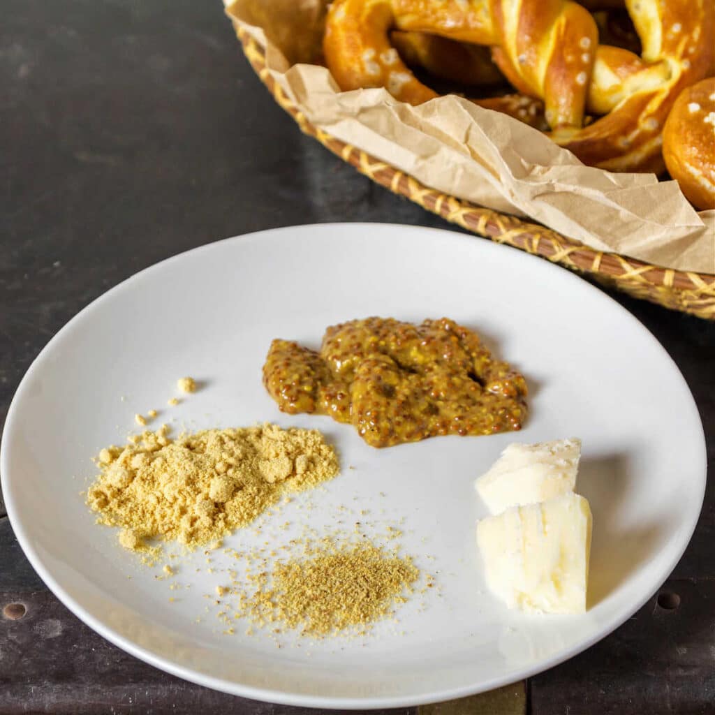 A white plate with dry mustard powder on it, white pepper, a chunk of cheese, and other spices. There is a corner of a basket of soft pretzels in the background.