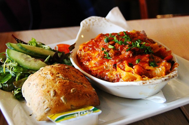 A single serving consisting of an oven proof-oval dish of baked pasta with Italian Sausage Pasta sauce over pasta, a dinner roll and tossed green salad all on a square white plate.