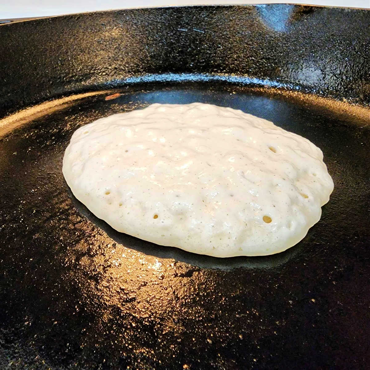 A closeup of pancake batter in a cast iron skillet. The batter is wet on top but set around the edges. This is how you can tell it is ready to flip.