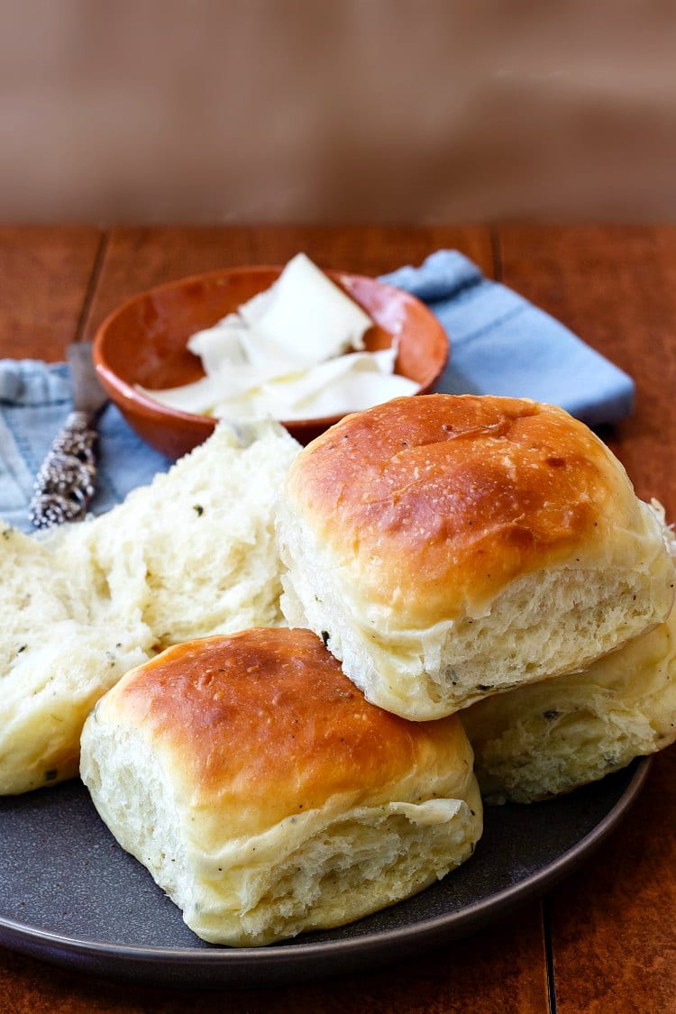 A stack of three soft dinner rolls on a gray plate with one split dinner roll and a bowl of butter in the background.