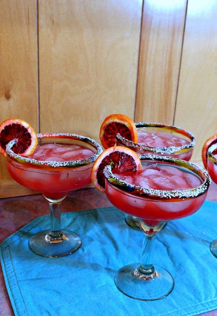 4 large cocktails in margarita glasses with salt on the rim and garnished with sliced blood oranges.