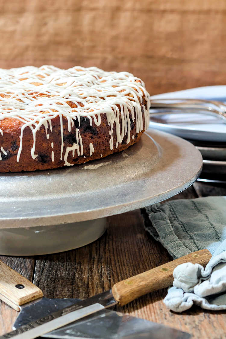 A whole brown butter blueberry buckle on a cake stand with a blue napkin and a stack of plates in the background.