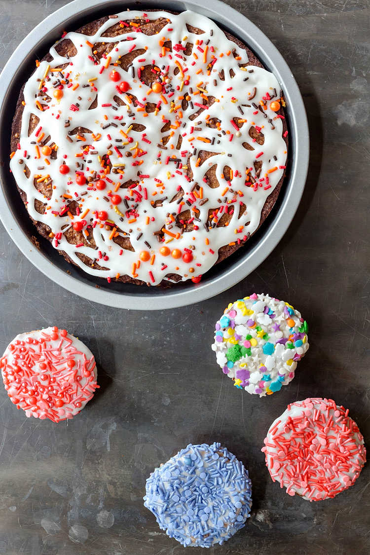 Overhead shot of a glazed cake in a round pan and four cupcakes covered in different colored sprinkles.