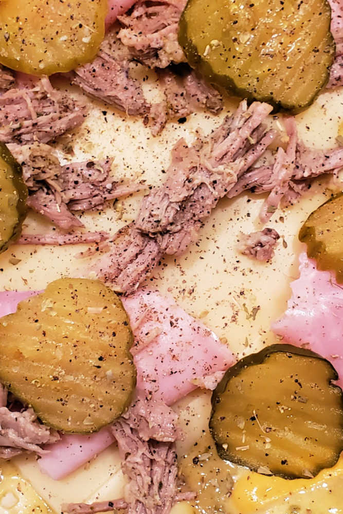 Close up of pickle chips, pulled pork, ham, and Swiss on pizza dough.