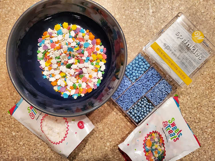 A bowl of multi colored sprinkles and bags of miscellaneous sprinkles.