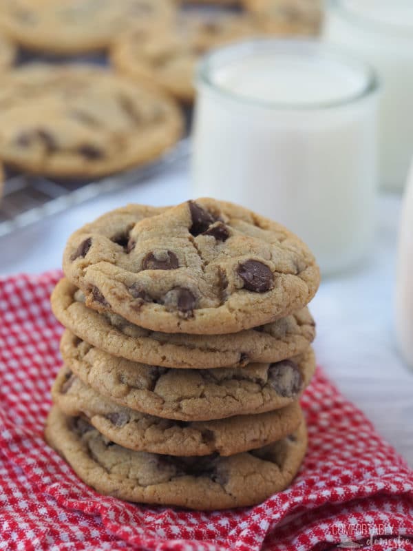 A stack of chocolate chip cookies on a towel with a glass of milk in the background.