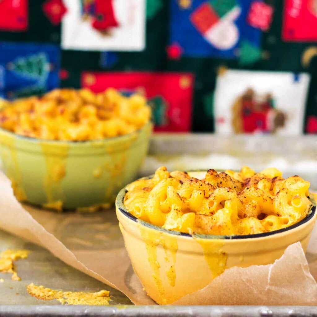 Two ramekins of gooey macaroni and cheese in front of a patchwork, Christmas-themed background.