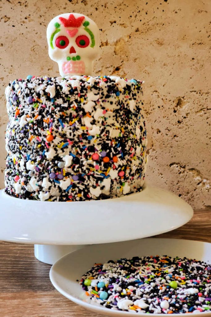 A day of the dead cake on a white platter with a sugar skull on top and a dish of sprinkles to the side.