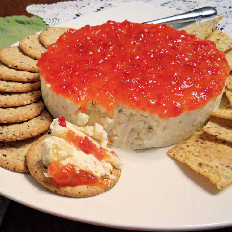 A savory cheesecake topped with pepper jelly spread over the top. A scoop has been taken out and smeared on a cracker. The cheesecake is surrounded with crackers on a white plate.