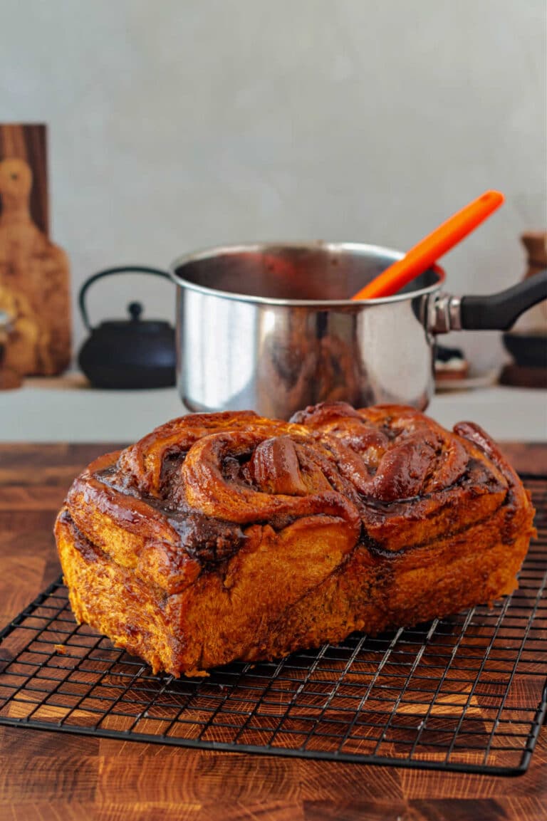 A whole pumpkin babka on a cooling rack with a small stainless steel pan with a silicone brush in it in the background.