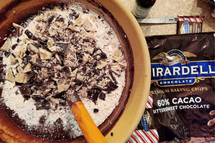 A bowl of brownie batter with flour and chopped peppermint bark in it next to a bag of Ghirardelli chocolate chips.