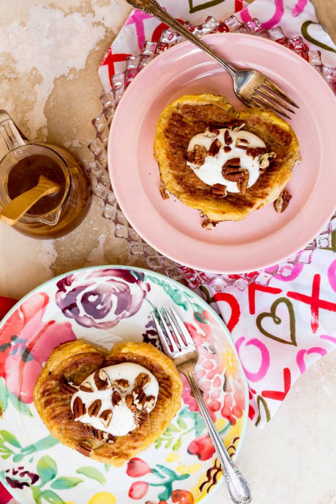 Overhead shot of 2 stuffed French toasts in the shape of hearts with cream and chopped pecans, one a floral plate and one on a pink plate.