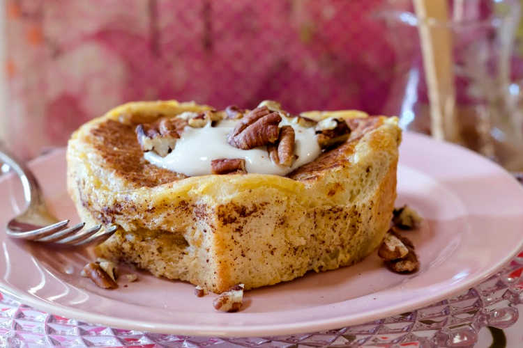 Side view of thick apple butterscotch french toast topped with brown sugar sweetened sour cream and toasted pecans on a pink plate with a fork.