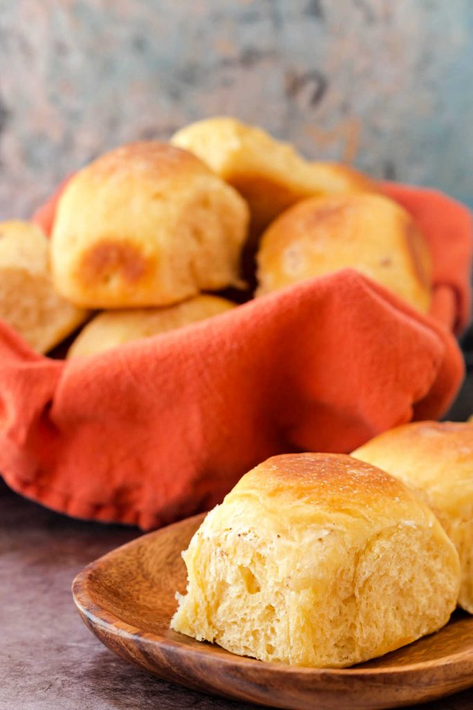 Basket of rolls with an orange napkin and a cheese roll on a wooden plate.