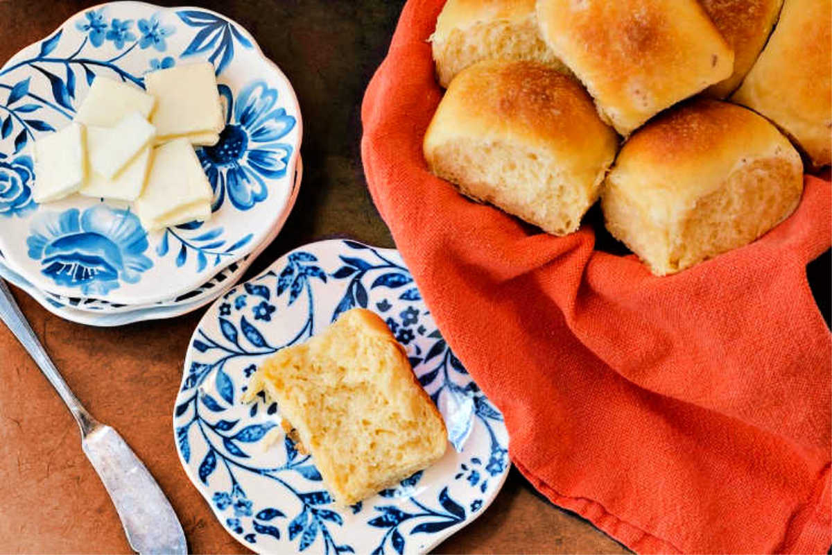 overhead shot of a basket of potato rolls, a plate with slices of butter, and a plate with a split cheese roll