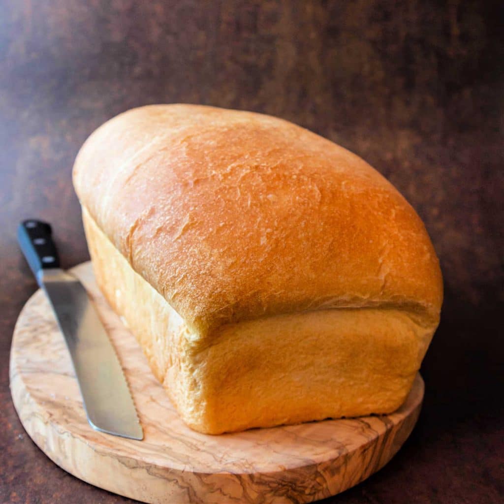A loaf of white sandwich bread on a board with a bread knife.