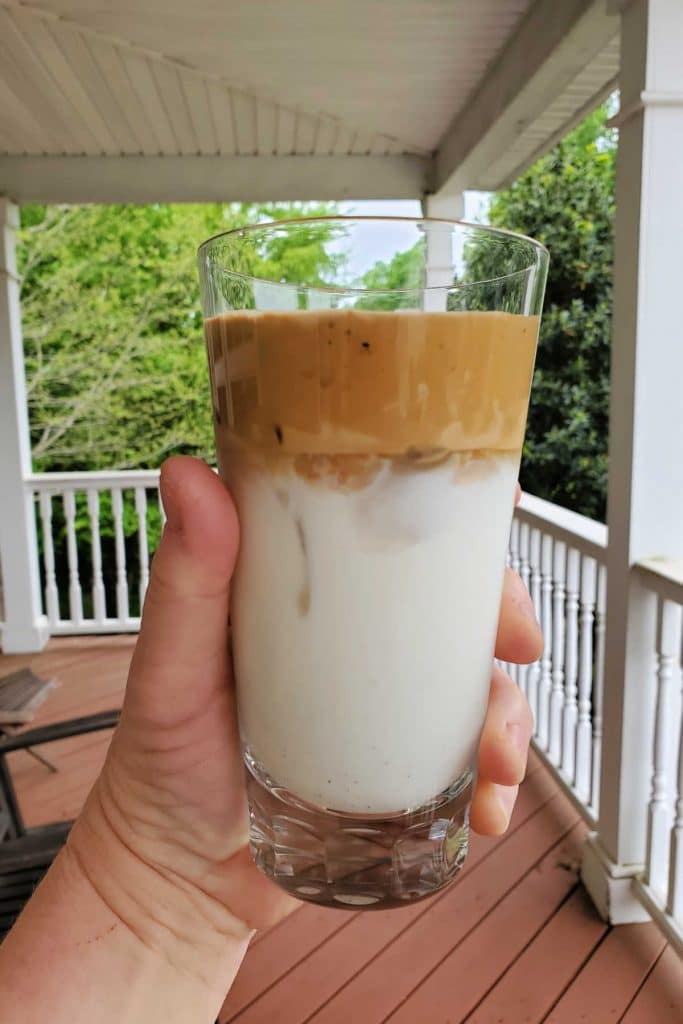 A hand holding a glass of dalgona coffee spooned on top of iced milk. The shot is outside on a porch in the summertime.