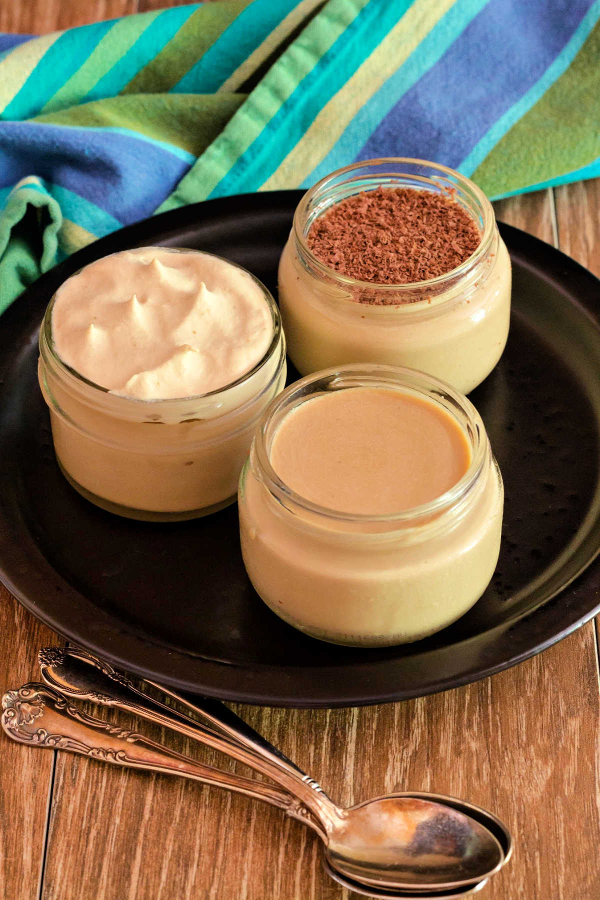 3 glass jars of coffee panna cotta on a black plate with 3 silvers spoons ready for serving.