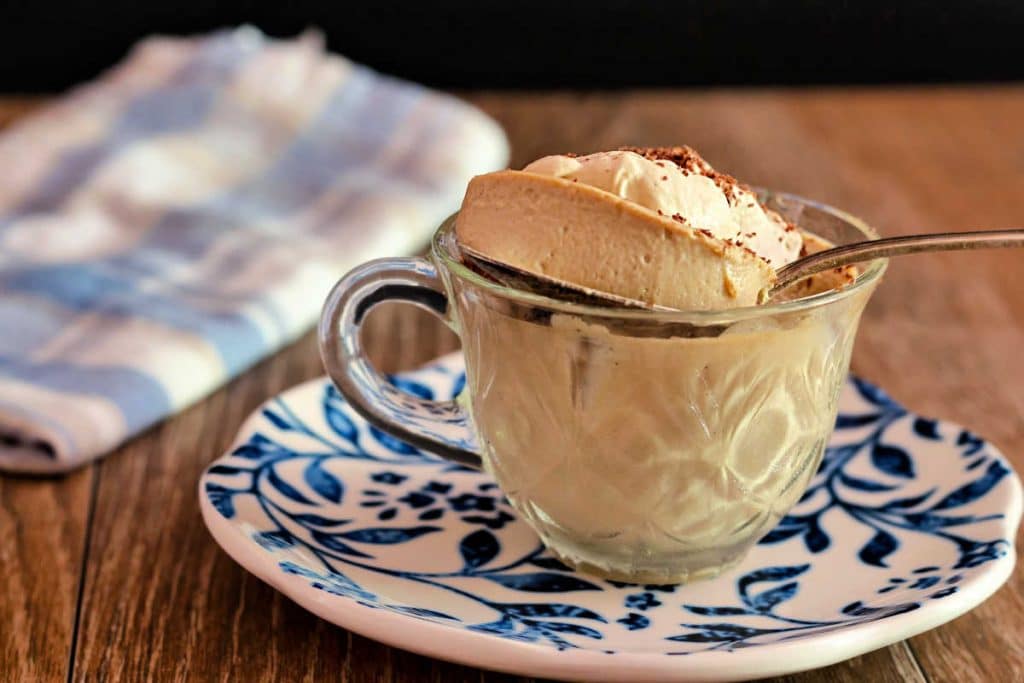 A bite of creamy beige Coffee panna cotta on a spoon coming out of a clear coffee cup on a blue and white plate.