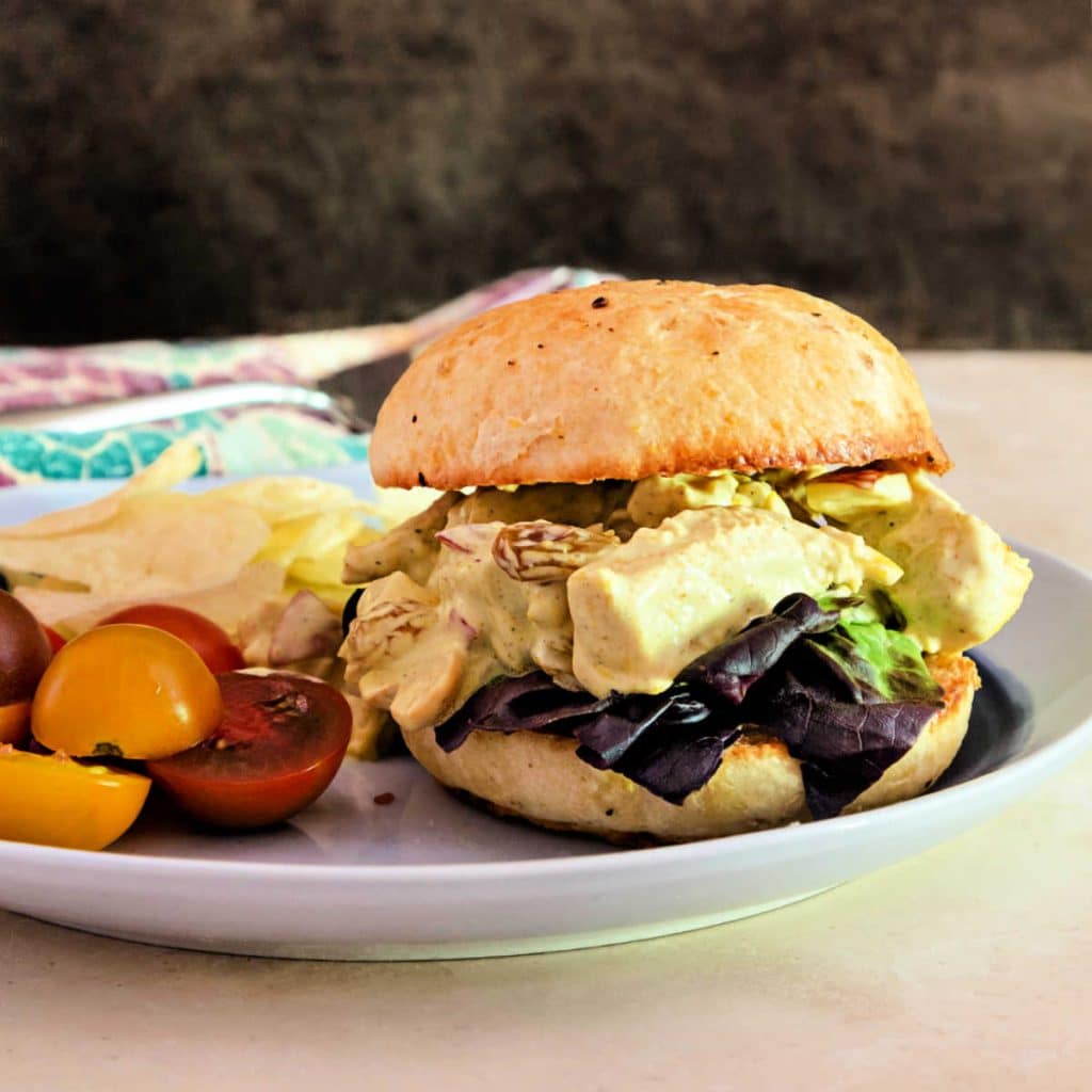 A sandwich of coronation chicken salad on a homemade bun with a garnish of cherry tomatoes and potato chips.