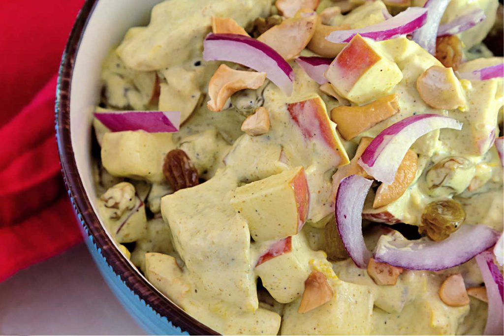 A bowl of curried chicken salad with obvious pieces of red onion, red apple, golden raisins, and cashew nuts.