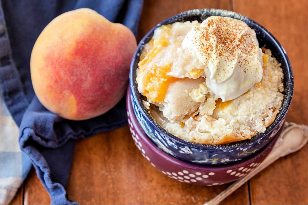 A  bowl of peach cobbler with whipped cream and sprinkle of cinnamon on top with a fresh peach next to it.