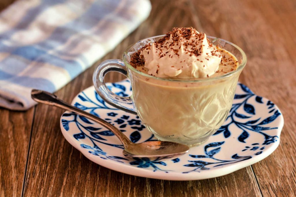 A clear coffee cup of panna cotta with whipped cream  and cocoa sprinkled on top on a blue and white plate.