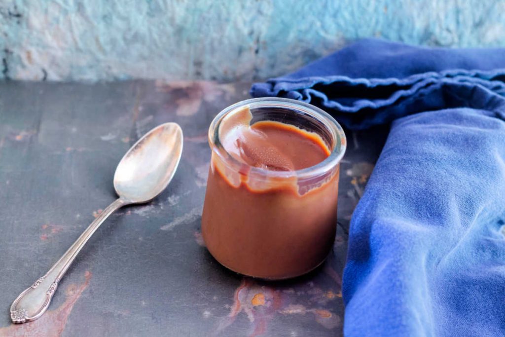 A jar of caramel chocolate pudding with silver spoon and blue napkin.