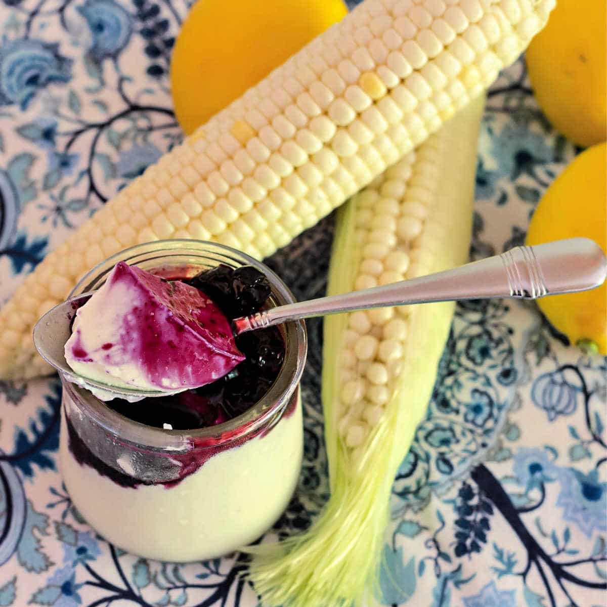 A glass of lemon corn panna cotta with blueberry sauce with lemons and ears of corn in the photo.