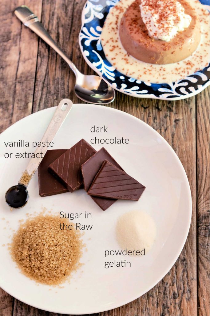 On a white plate, the panna cotta ingredients laid out, with text next to each. A metal measuring spoon with vanilla paste 9or vanilla extract) in it, squares of dark chocolate, a small mound of dry powdered gelatin, a mound of brown sugar in the raw granules.