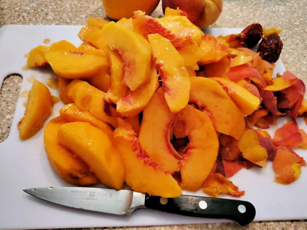 A cutting board with a pile of fresh peaches peeled and sliced, their pits to the side and a paring knife in front.