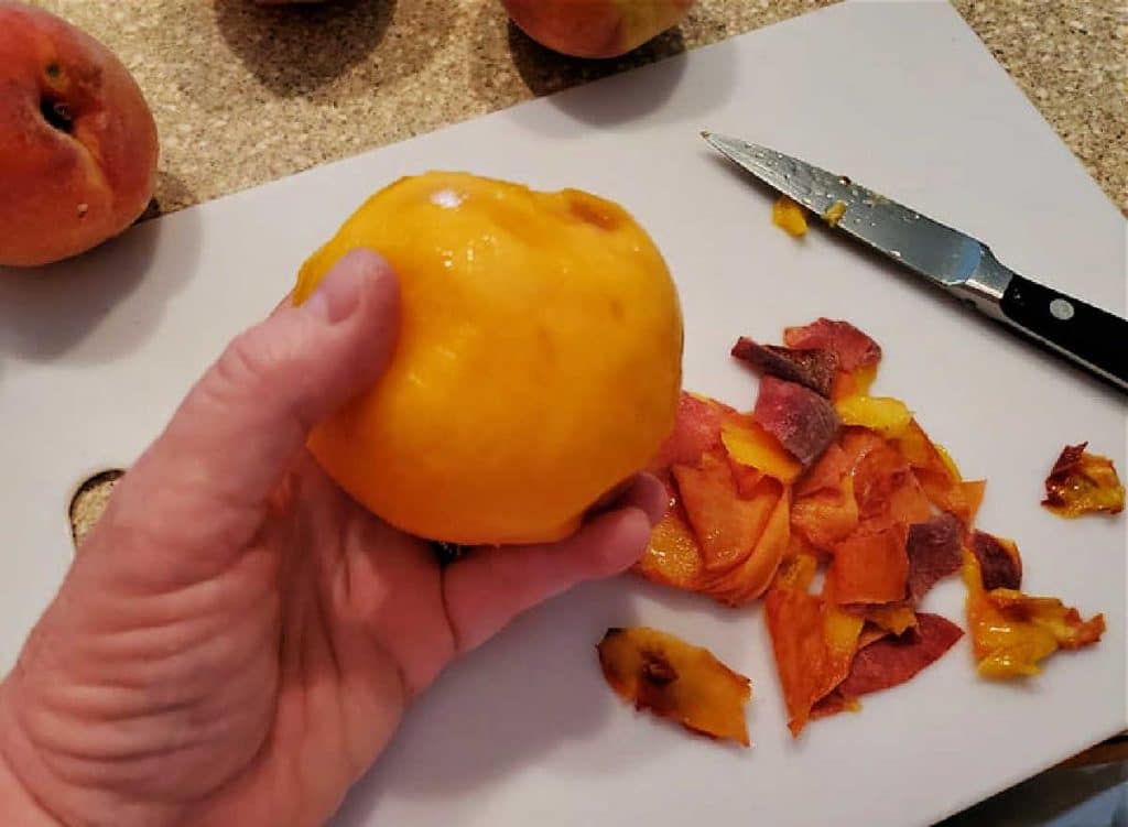 A hand holding a peeled peach with peelings and a paring knife on a cutting board.