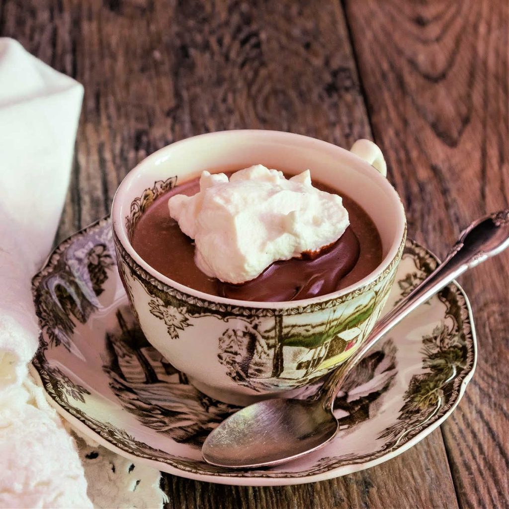 A vintage teacup decorated with brown and green scenes of farms and trees. Cup is filled with chocolate panna cotta with whipped cream on top. A silver spoon rests on the decorated saucer.